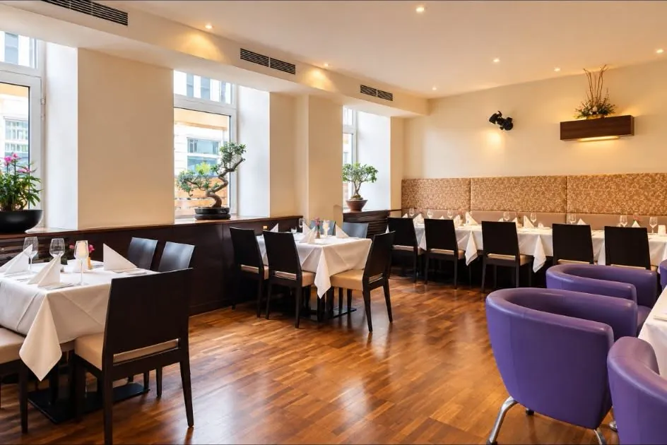 Heller Gastraum mit eingedeckten Tischen, Bonsai an den Fenstern und violetten Lounge-Sesseln