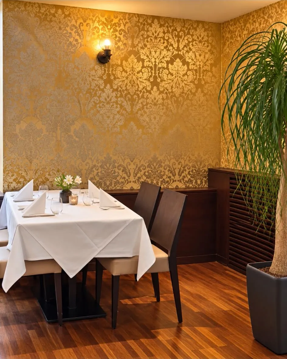 Eingedeckter Tisch vor der goldenen Ornamenttapete, daneben eine Zimmerpflanze
